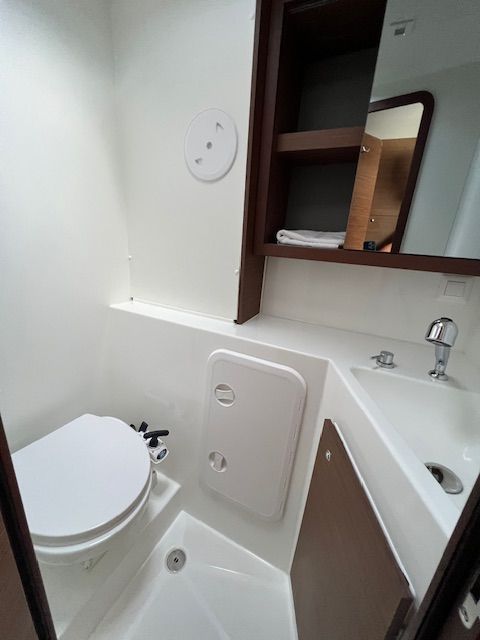Lagoon 42 2021 Salle de bain cabine avant Petite salle de bain avec toilette, lavabo et miroir. Mur blanc et rangement en bois.
