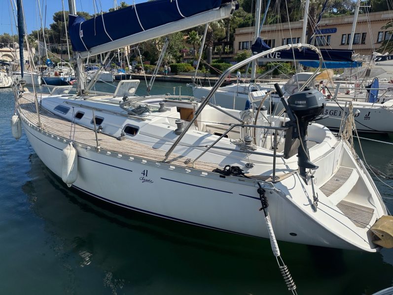 Grand Sud 55 Voilier blanc accosté dans un port, avec d'autres bateaux en arrière-plan. Ciel nuageux.