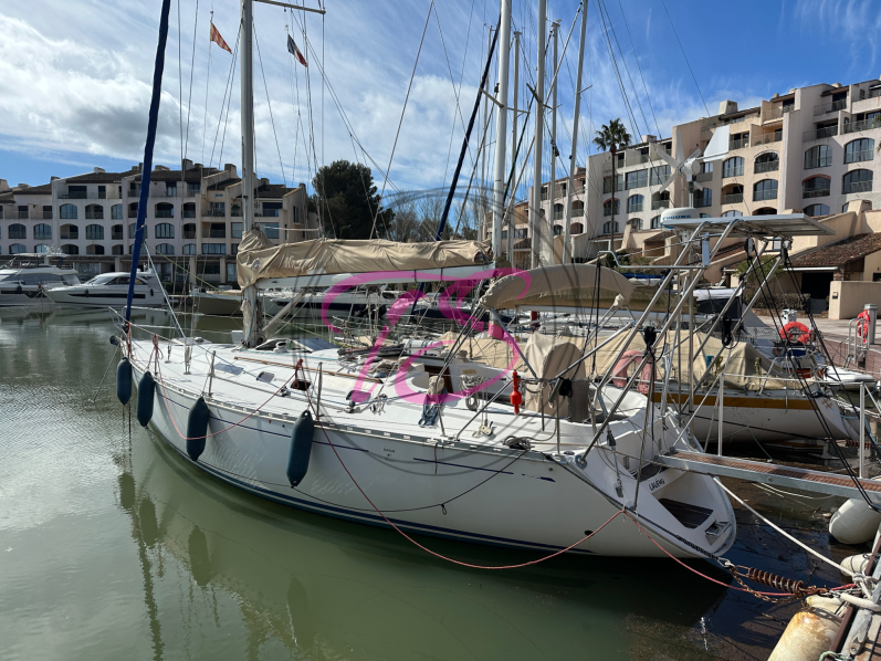 Voilier blanc accosté dans un port, avec d'autres bateaux en arrière-plan. Ciel nuageux.