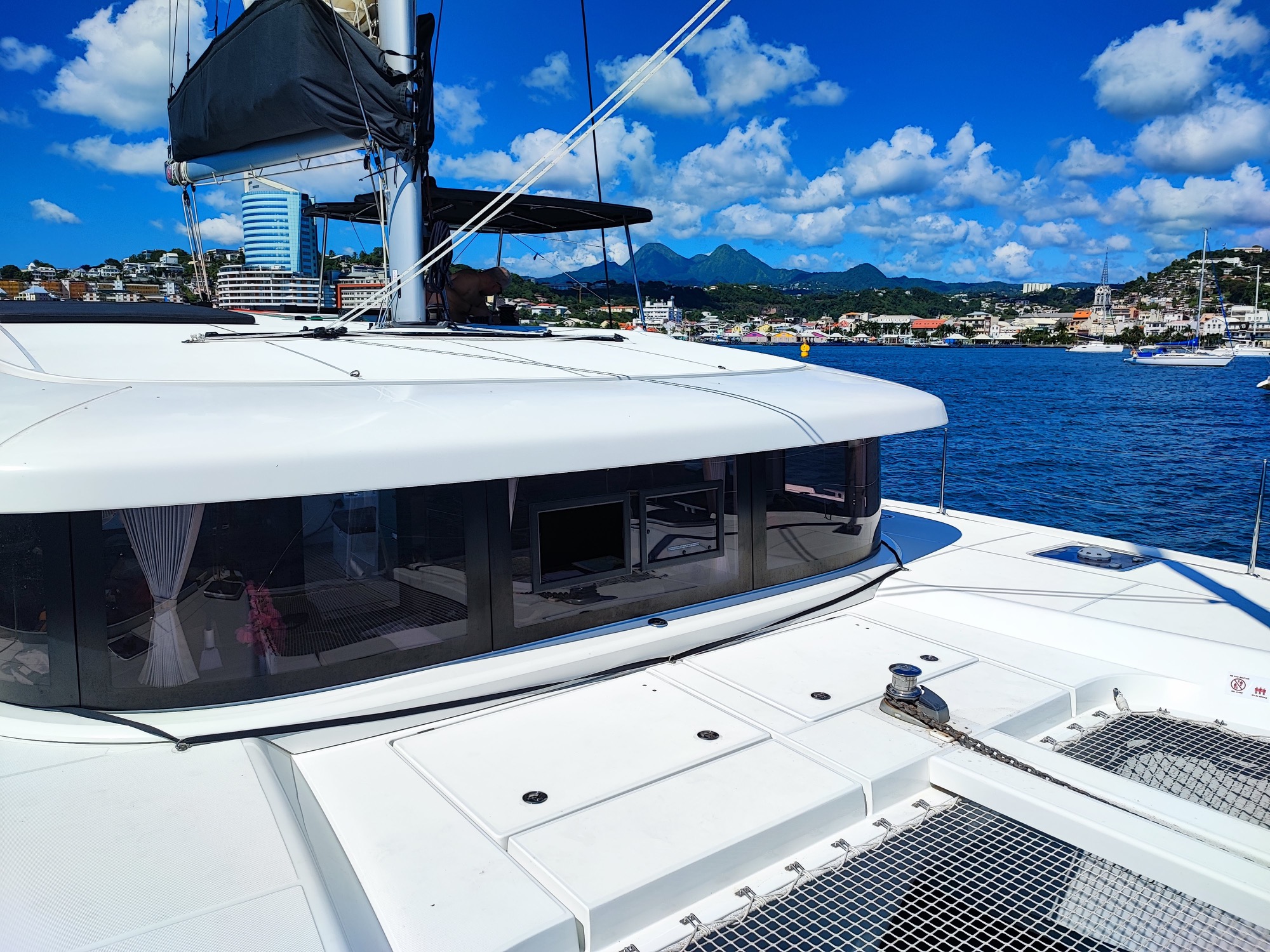 Lagoon 42 2021 Pont avant Vue d'un catamaran blanc au port, avec des montagnes et des bateaux en arrière-plan.