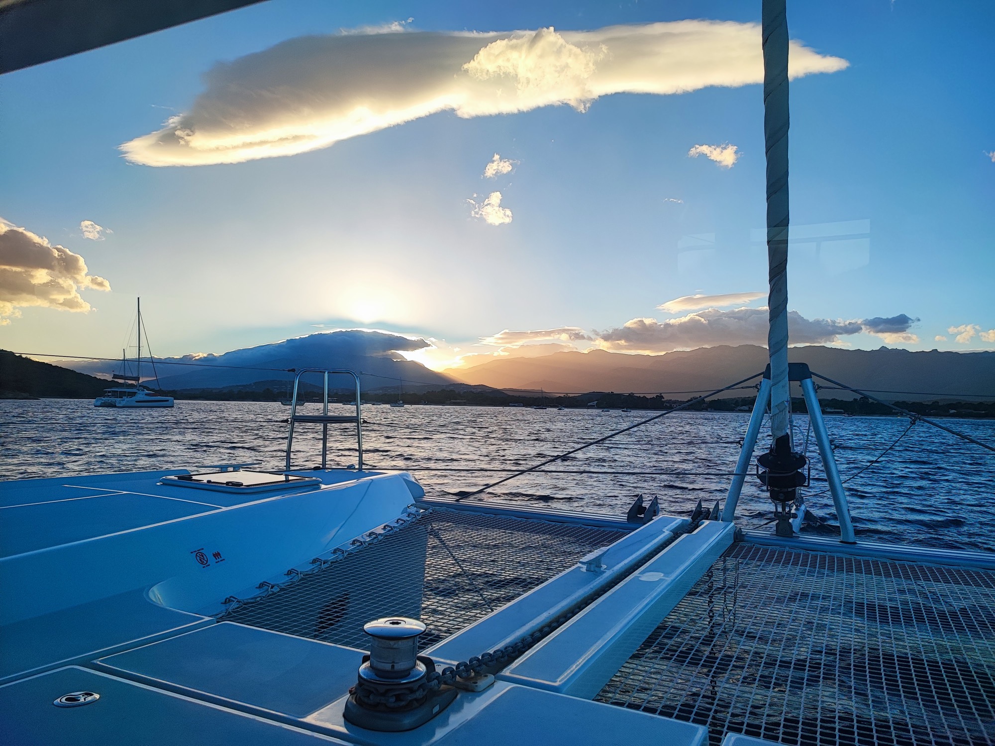 Lagoon 42 2021 Pont avant Coucher de soleil sur la mer vue depuis un bateau, avec des nuages et des montagnes en arrière-plan.
