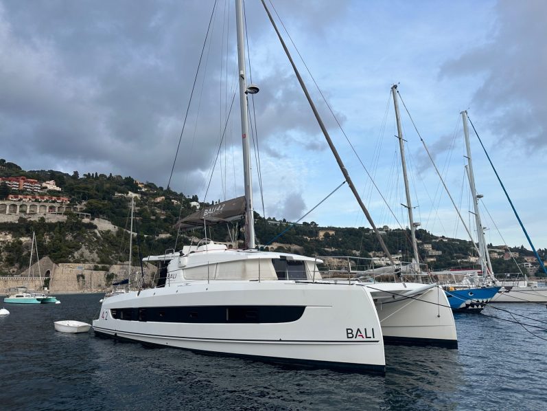 Voilier blanc accosté dans un port, avec d'autres bateaux en arrière-plan. Ciel nuageux.